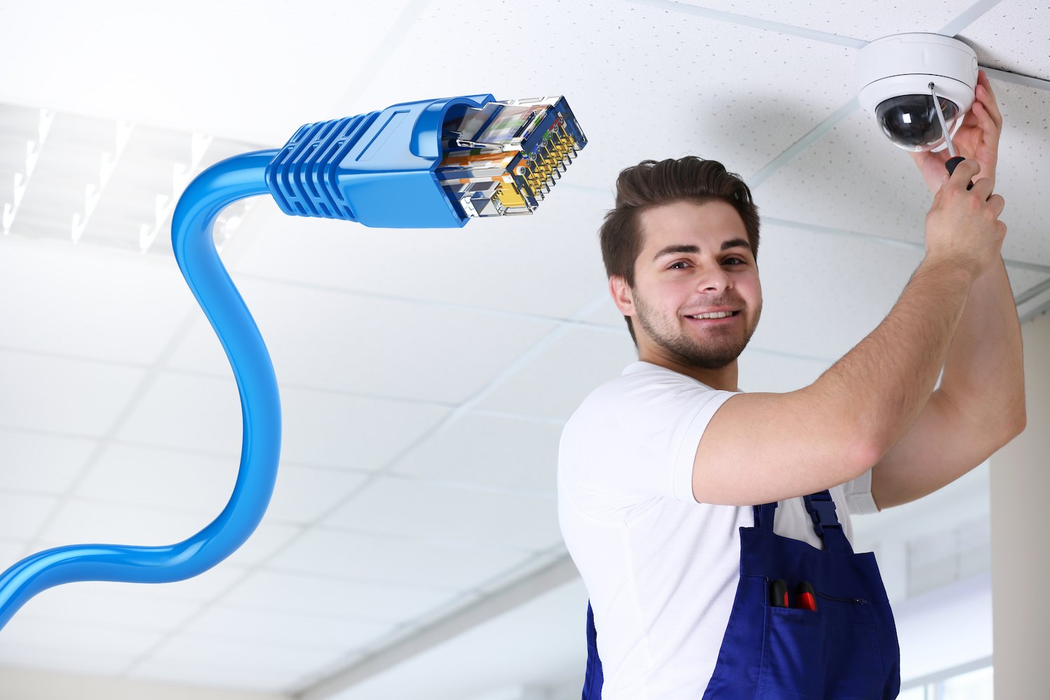 A workman in overalls installs an overhead camera on a white ceiling. Shown in the foreground is a blue Ethernet cable.