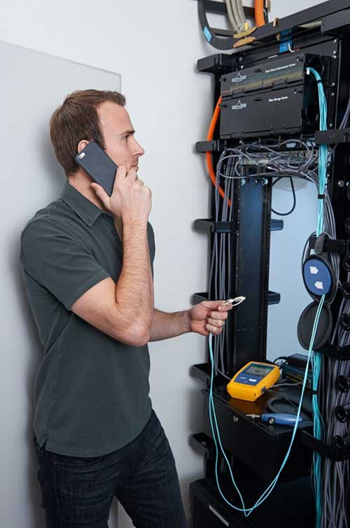 Image of a technician on the phone in a telecommunications room using an OTDR to troubleshoot a fiber link